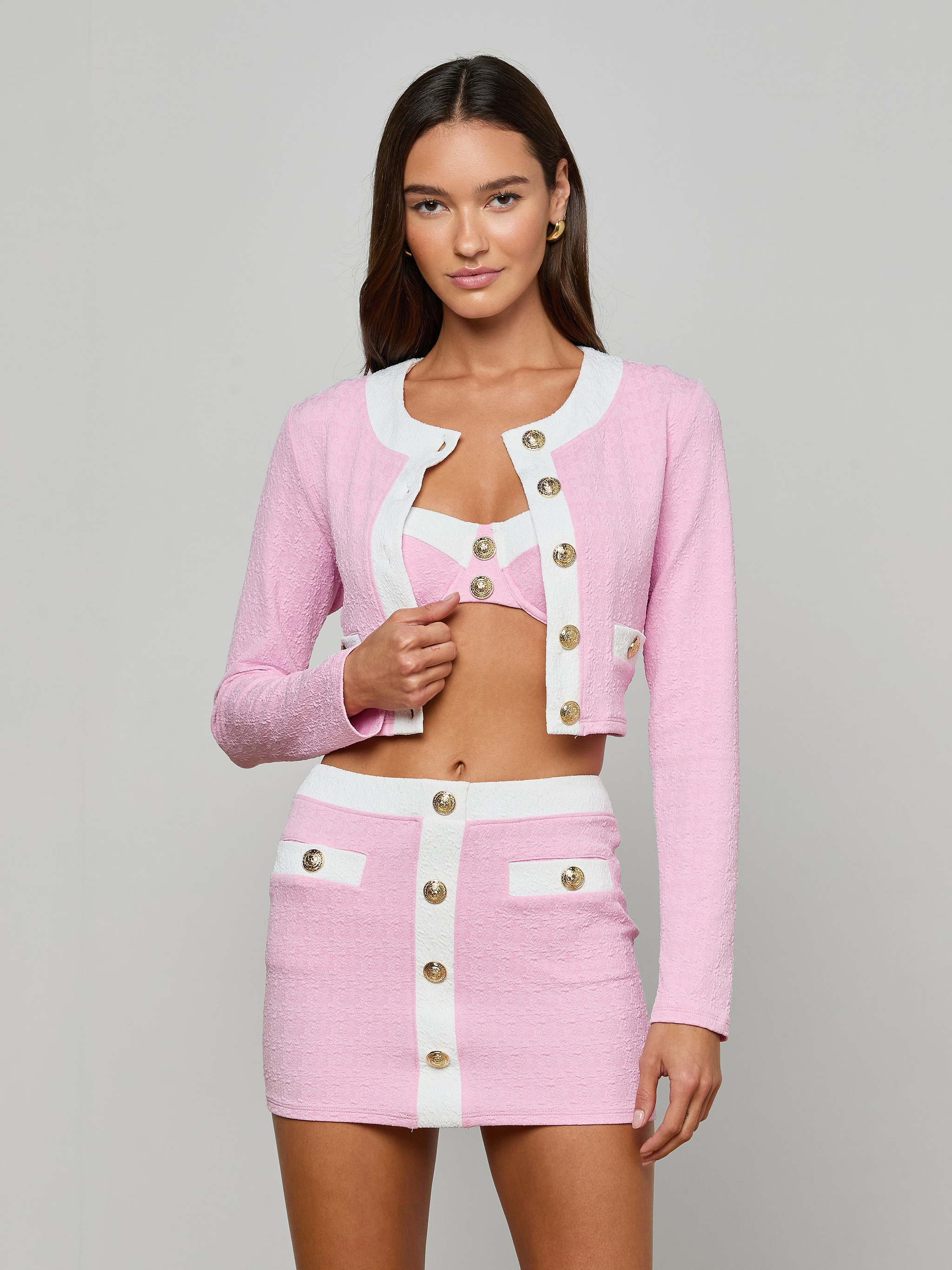 A woman stands against a plain background wearing the Adaline Cardigan Cover-Up, a pink and white textured bouclé set with gold buttons. The look features a cropped jacket, matching bralette, and a high-waisted mini skirt with white trim and pocket details.