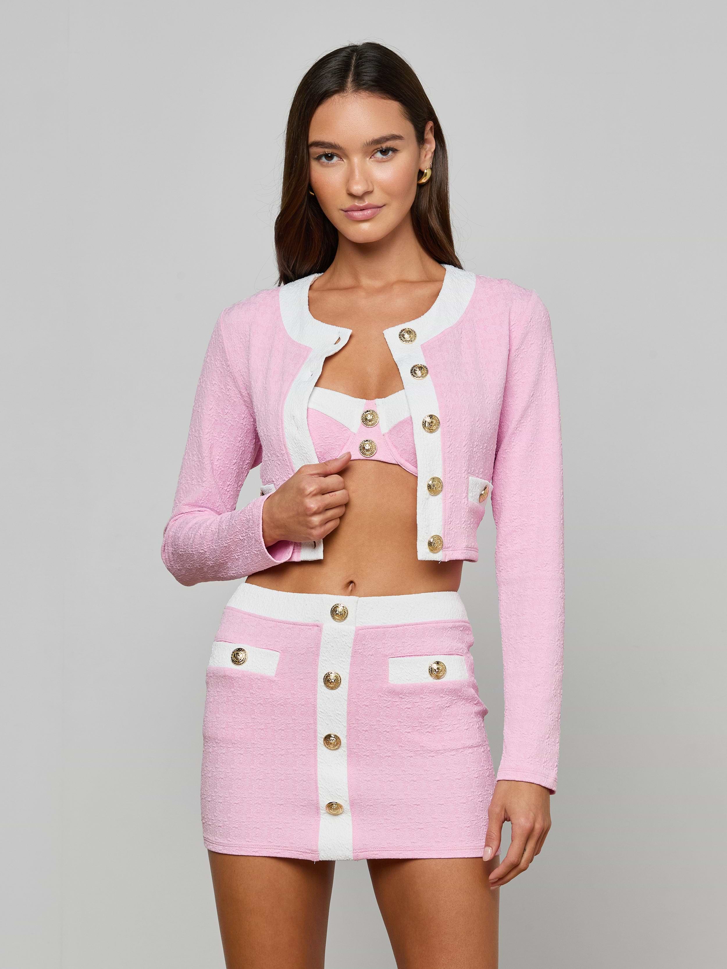 A woman stands against a plain background wearing the Adaline Cardigan Cover-Up, a pink and white textured bouclé set with gold buttons. The look features a cropped jacket, matching bralette, and a high-waisted mini skirt with white trim and pocket details.