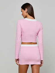 A woman faces away, wearing the Adaline Cardigan Cover-Up— a light pink textured bouclé long-sleeve crop top and matching high-waisted mini skirt with white trim, set against a plain gray background.