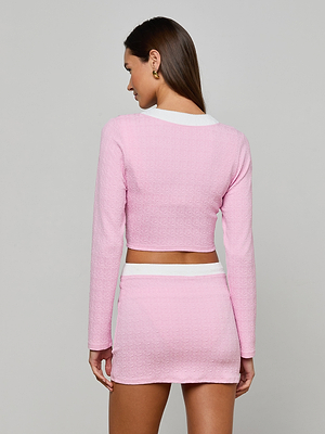A woman faces away, wearing the Adaline Cardigan Cover-Up— a light pink textured bouclé long-sleeve crop top and matching high-waisted mini skirt with white trim, set against a plain gray background.