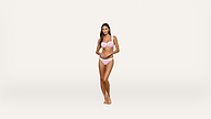 A woman stands barefoot against a plain, light background, wearing a light pink bikini and looking toward the camera with a neutral expression.
