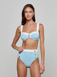 A woman stands against a plain, light background, looking at the camera with a neutral expression while wearing the Vanessa High-Waisted Bikini Bottom in light blue and white bouclé swim fabric, featuring decorative buttons and a supportive fit.