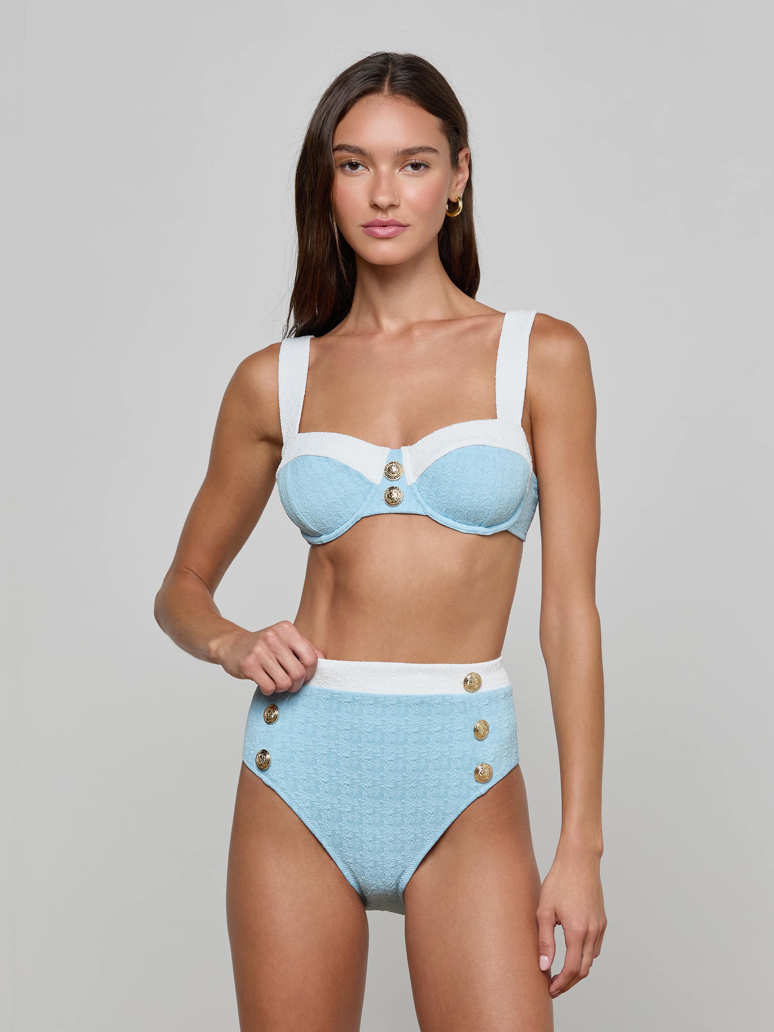 A woman stands against a plain, light background, looking at the camera with a neutral expression while wearing the Vanessa High-Waisted Bikini Bottom in light blue and white bouclé swim fabric, featuring decorative buttons and a supportive fit.