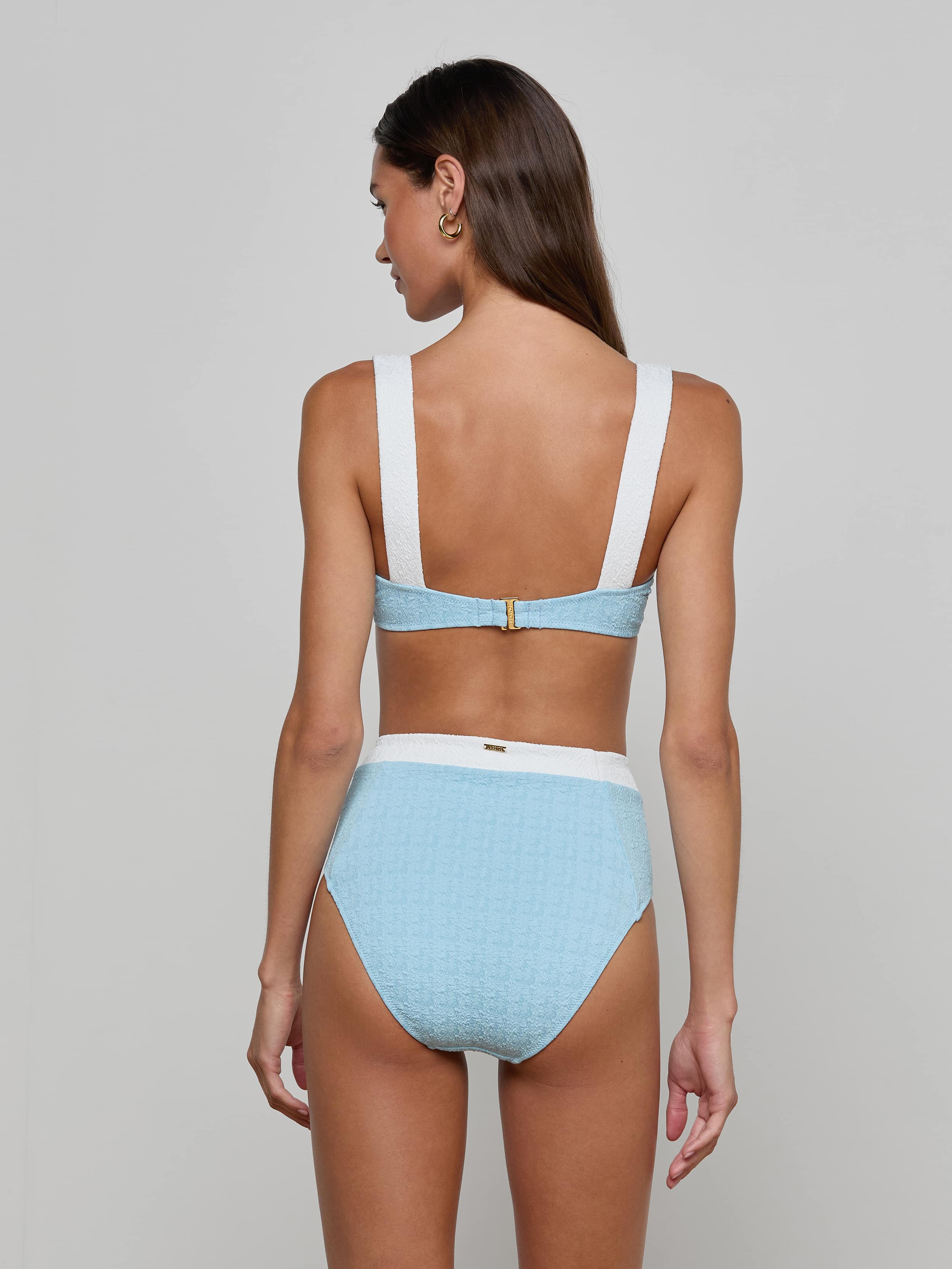 A woman with long brown hair models the Vanessa High-Waisted Bikini Bottom in light blue bouclé swim fabric with white straps and waistband, shown from the back against a plain light background. This supportive bikini bottom offers moderate coverage.