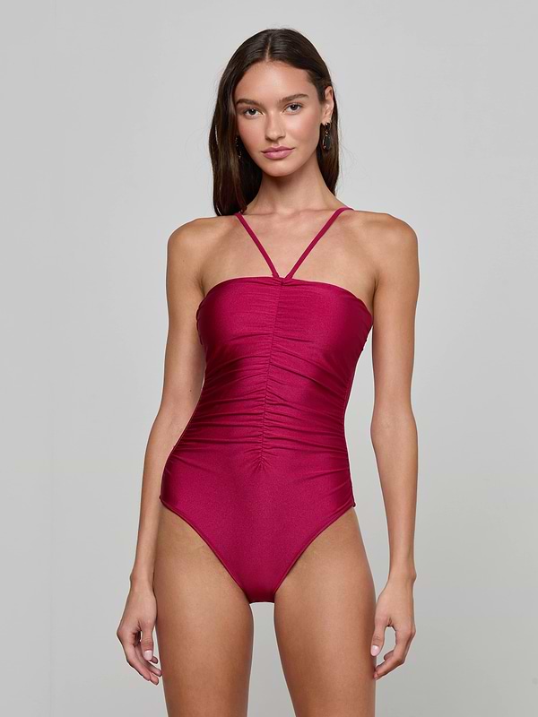 A woman stands wearing the Aubrey Ruched One-Piece Swimsuit, a sleeveless burgundy design featuring a high-cut leg, thin straps, and textured shirring. The background is plain and light-colored.