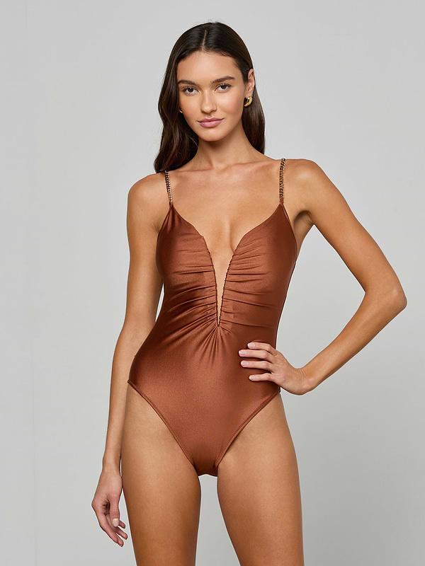 A woman models the Lilou One-Piece Swimsuit—a shiny brown swimsuit featuring a deep v-wire plunge neckline, thin straps, and a high-leg cut—as she stands with one hand on her hip against a plain light background.