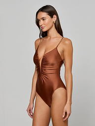 A woman stands against a plain light background, wearing the Lilou One-Piece Swimsuit in a shiny, deep brown shade. This swimsuit features a plunging v-wire neckline, thin straps, and a high-leg cut that accentuates her silhouette.