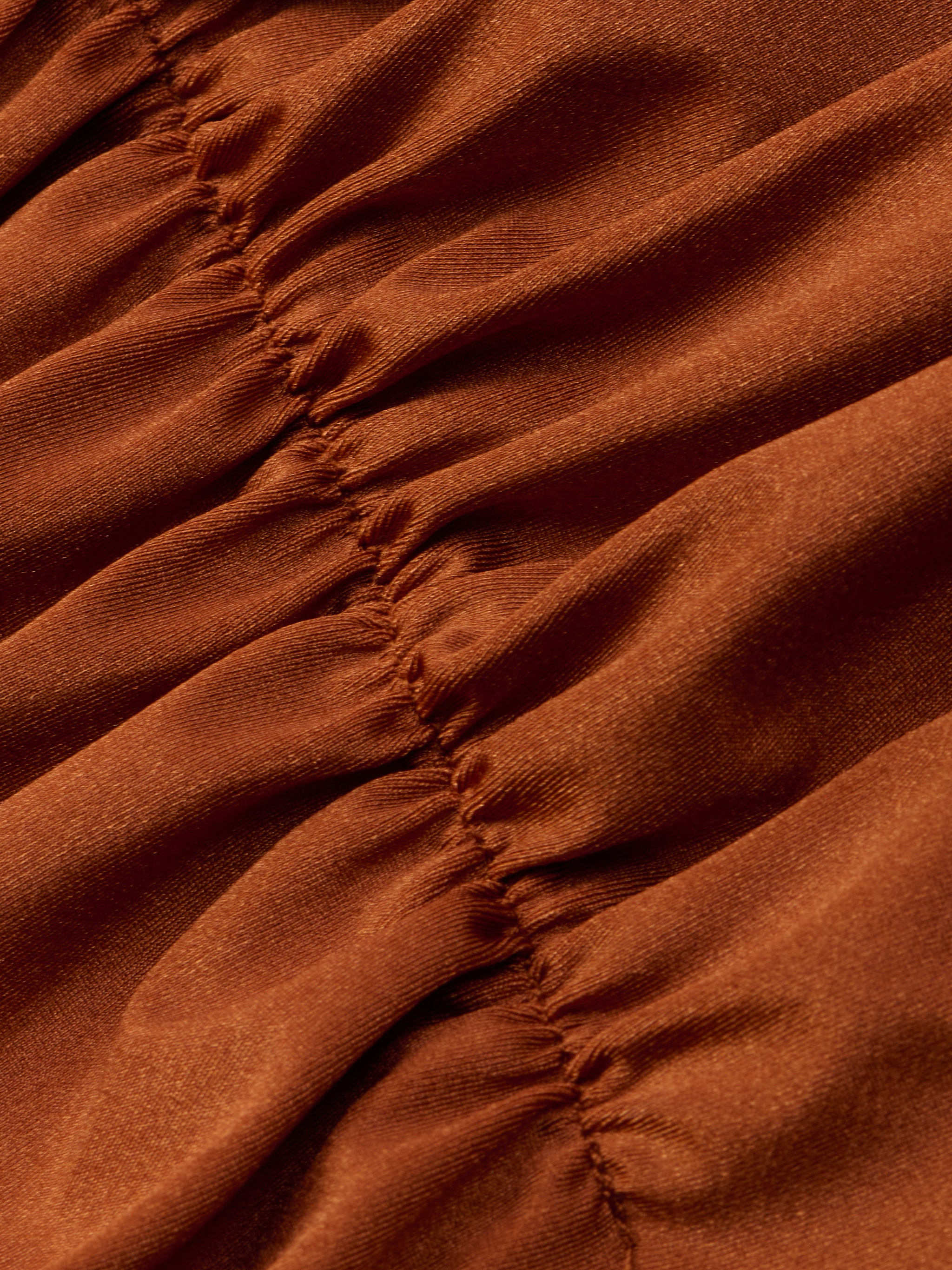 A close-up of the Lilou One-Piece Swimsuit in rust shows gathered fabric with visible vertical stitching that creates soft folds and textured, ruched lines. The material looks smooth and slightly shiny, highlighting the high-leg cut design.