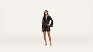 A woman stands barefoot on a plain white background, wearing a long-sleeved, black lace dress with a v-neckline and a short hem. She has one hand on her hip and a neutral expression.