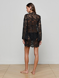 A woman stands barefoot with her back to the camera, wearing the Ansley Cover-Up Dress—a black, long-sleeve paisley crochet design that is semi-transparent. The background is plain and light-colored.