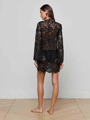 A woman stands barefoot on a wooden floor, facing away, wearing the black Ansley Cover-Up Dress by L'AGENCE with sheer paisley crochet long sleeves and a visible black undergarment, set against a plain light-colored wall.