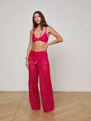 A woman stands indoors on a wooden floor, posing in a bright pink bikini top and the Geraldine Cover-Up Pant—sheer paisley lace pants with a wide leg fit. She rests one hand on her hip against a plain white wall.