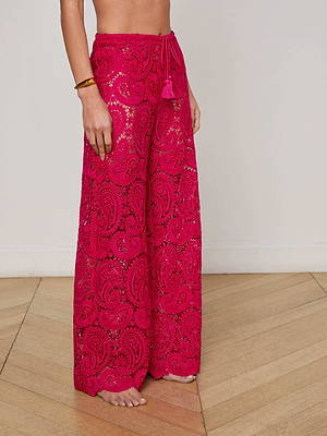 A person stands barefoot on a wooden floor, showing only their lower body dressed in the bright pink Geraldine Cover-Up Pant featuring sheer paisley lace, wide legs, and a drawstring waist.