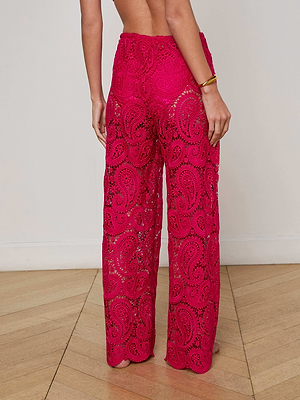 A woman stands barefoot on a wooden floor, back to camera, wearing bright pink Geraldine Cover-Up Pants with wide legs and sheer paisley lace. She accessorizes with a gold bracelet on her right wrist.