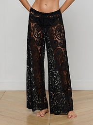 Wearing the Geraldine Cover-Up Pant—black, sheer wide-leg pants with a detailed paisley crochet lace design—the person stands barefoot, their upper body mostly out of frame against a plain background.