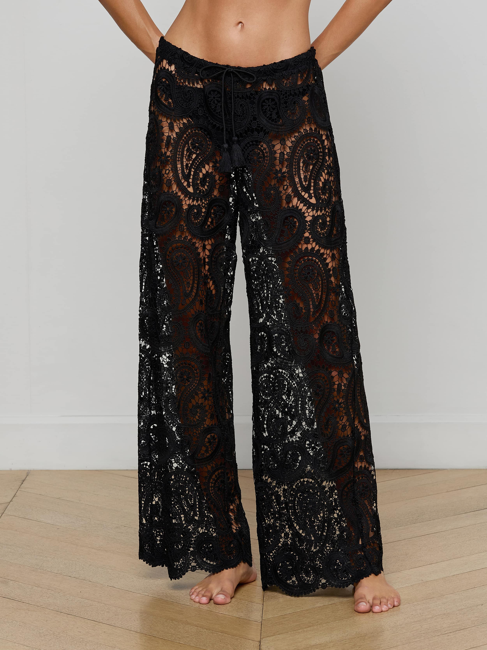 Wearing the Geraldine Cover-Up Pant—black, sheer wide-leg pants with a detailed paisley crochet lace design—the person stands barefoot, their upper body mostly out of frame against a plain background.