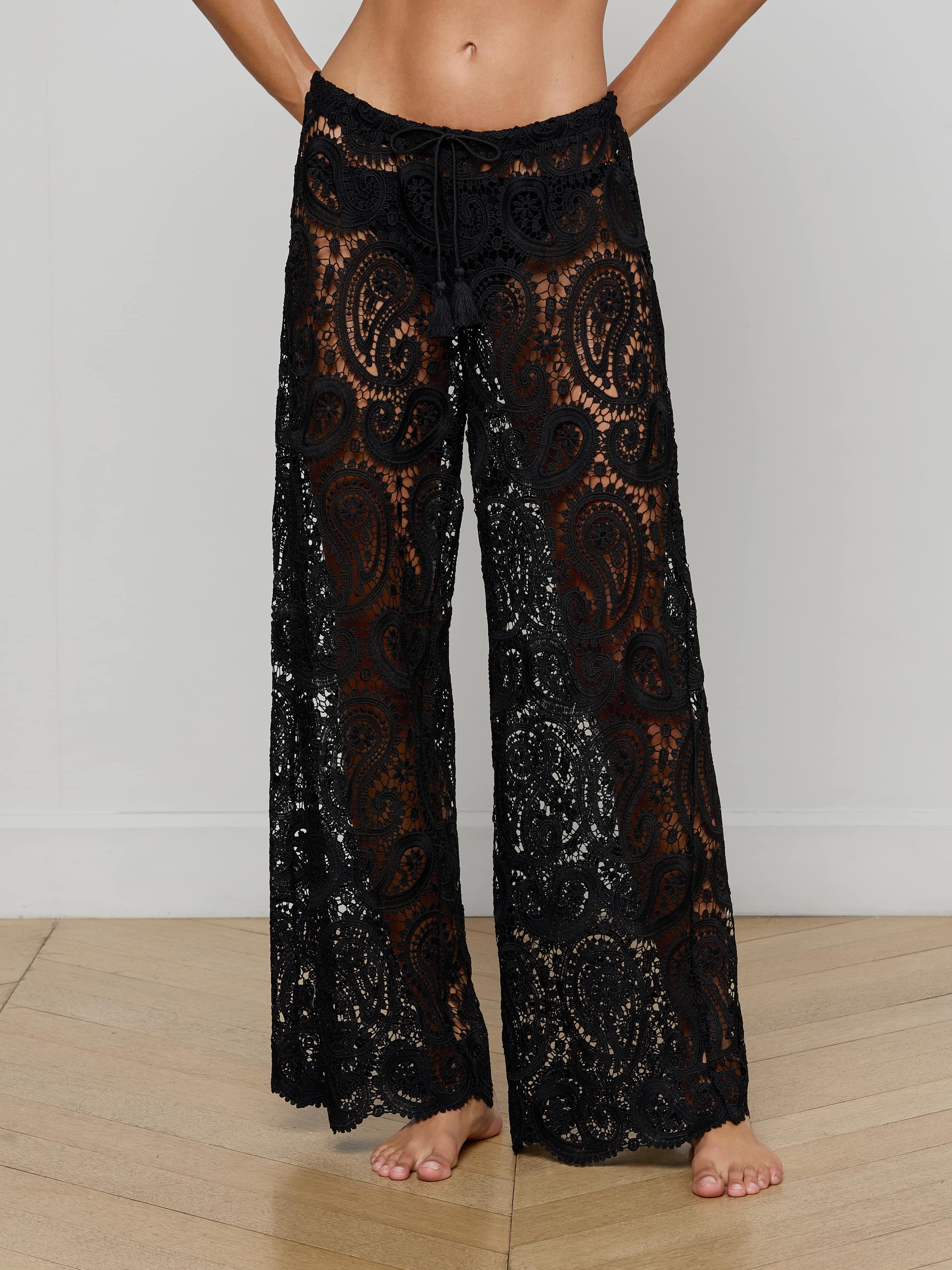 Wearing the Geraldine Cover-Up Pant—black, sheer wide-leg pants with a detailed paisley crochet lace design—the person stands barefoot, their upper body mostly out of frame against a plain background.