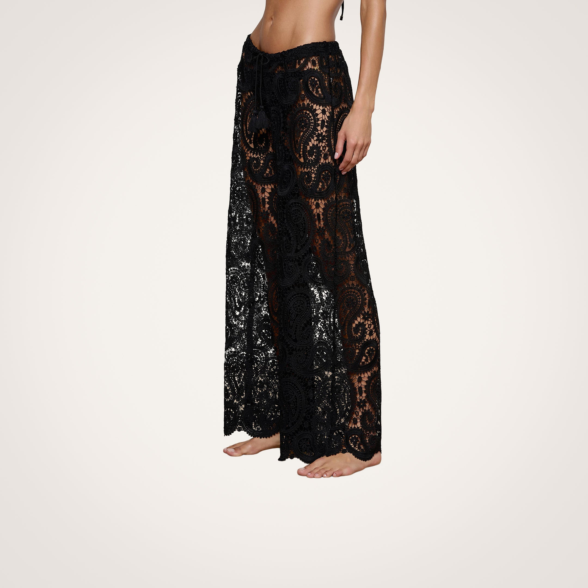 A woman stands barefoot, wearing the Geraldine Cover-Up Pant—black lace, see-through wide-leg bohemian pants. Her upper body is cropped out of the image, and the background is plain and light-colored.