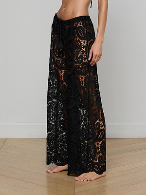 A person stands barefoot on a wooden floor, wearing the Geraldine Cover-Up Pant—black paisley crochet with a sheer lace design and drawstring waist, perfect for resort wear. Image is cropped from the chest down.