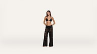 A woman stands barefoot against a plain white background, wearing a black bikini top and black sheer lace pants with a floral pattern. Her hands are clasped behind her back, and she faces forward.