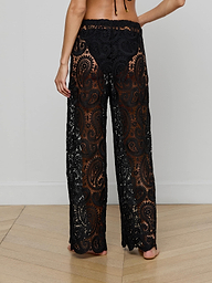 A person stands barefoot, seen from behind, wearing the Geraldine Cover-Up Pant—bohemian-style pants crafted from black lace with a paisley crochet pattern. The upper body is mostly out of frame against a plain, light-colored background.