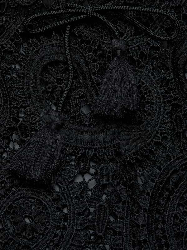 Close-up of the Geraldine Cover-Up Pant, featuring intricate black lace fabric adorned with decorative loops, a braided tie, and two tassels resting on top, creating a textured and elegant pattern reminiscent of resort wear.