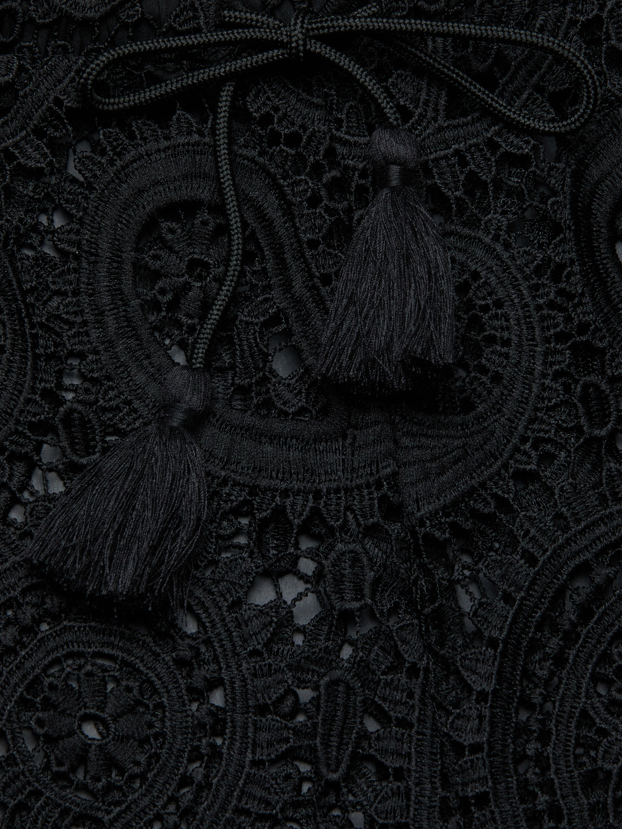 Close-up of the Geraldine Cover-Up Pant, featuring intricate black lace fabric adorned with decorative loops, a braided tie, and two tassels resting on top, creating a textured and elegant pattern reminiscent of resort wear.
