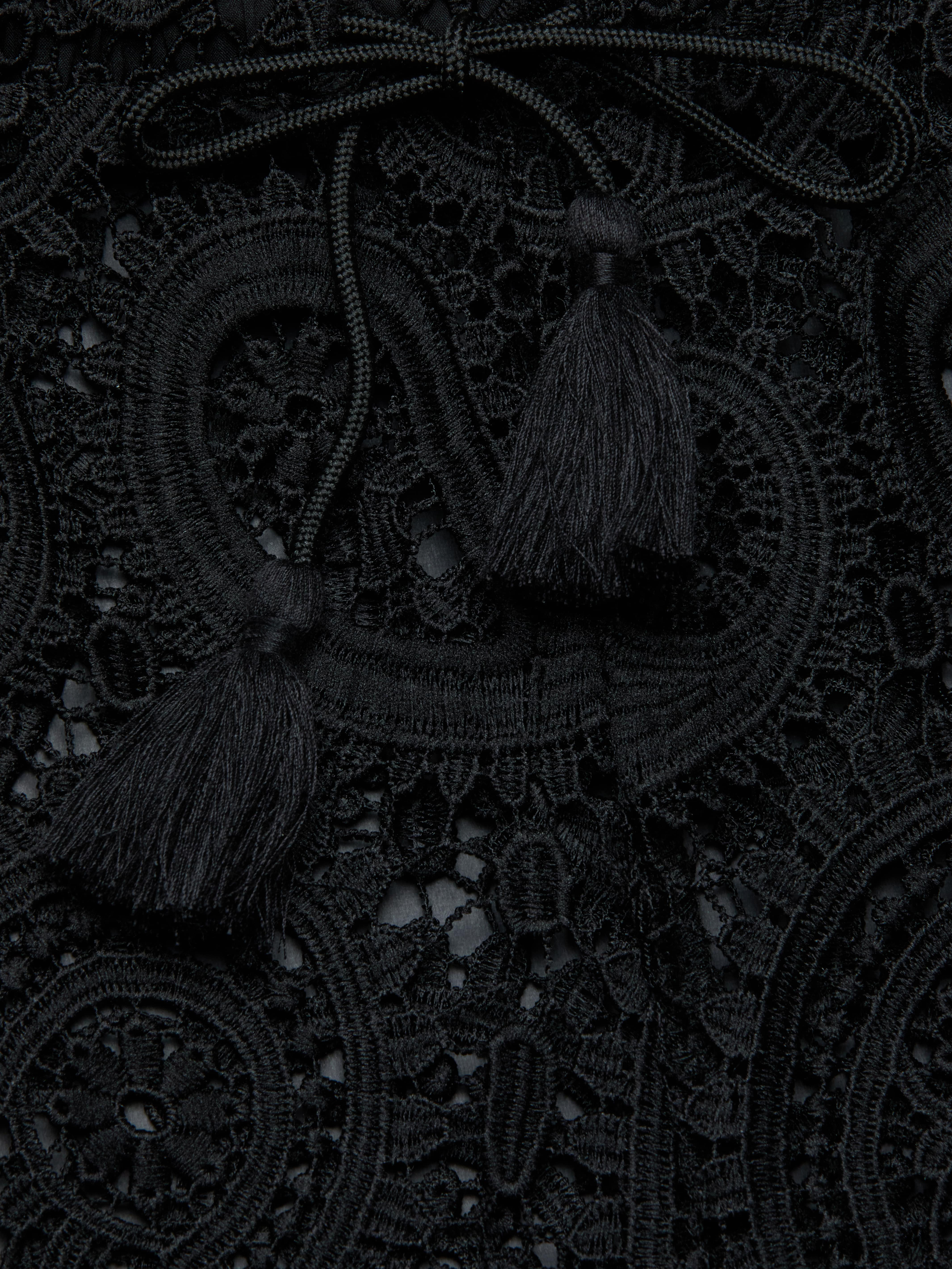 Close-up of the Geraldine Cover-Up Pant, featuring intricate black lace fabric adorned with decorative loops, a braided tie, and two tassels resting on top, creating a textured and elegant pattern reminiscent of resort wear.