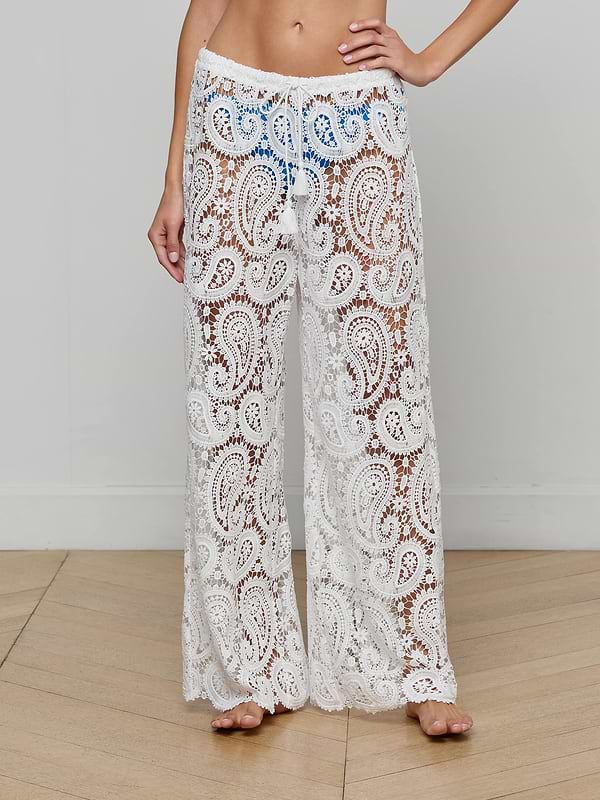 A person is modeling the Geraldine Cover-Up Pant—sheer, white bohemian pants featuring a paisley lace pattern and a loose fit with a drawstring waist, perfect for resort wear. The model is barefoot against a plain background.