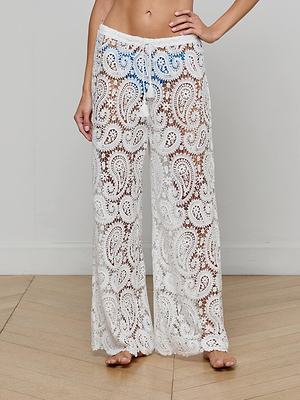 A person stands barefoot on a wooden floor, wearing the Geraldine Cover-Up Pant. These white, semi-transparent lace pants feature a paisley crochet pattern, drawstring waist, and subtly reveal blue undergarments. The person's face is not shown.
