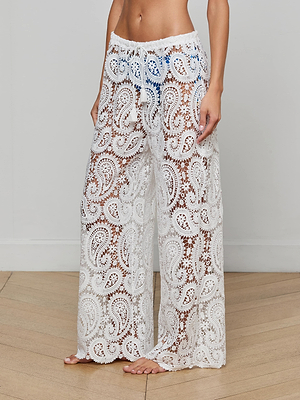 A person stands barefoot on a wooden floor wearing Geraldine Cover-Up Pants—white, sheer, wide-leg bohemian crochet with an adjustable drawstring waist. The semi-transparent resort wear reveals a blue bikini underneath.