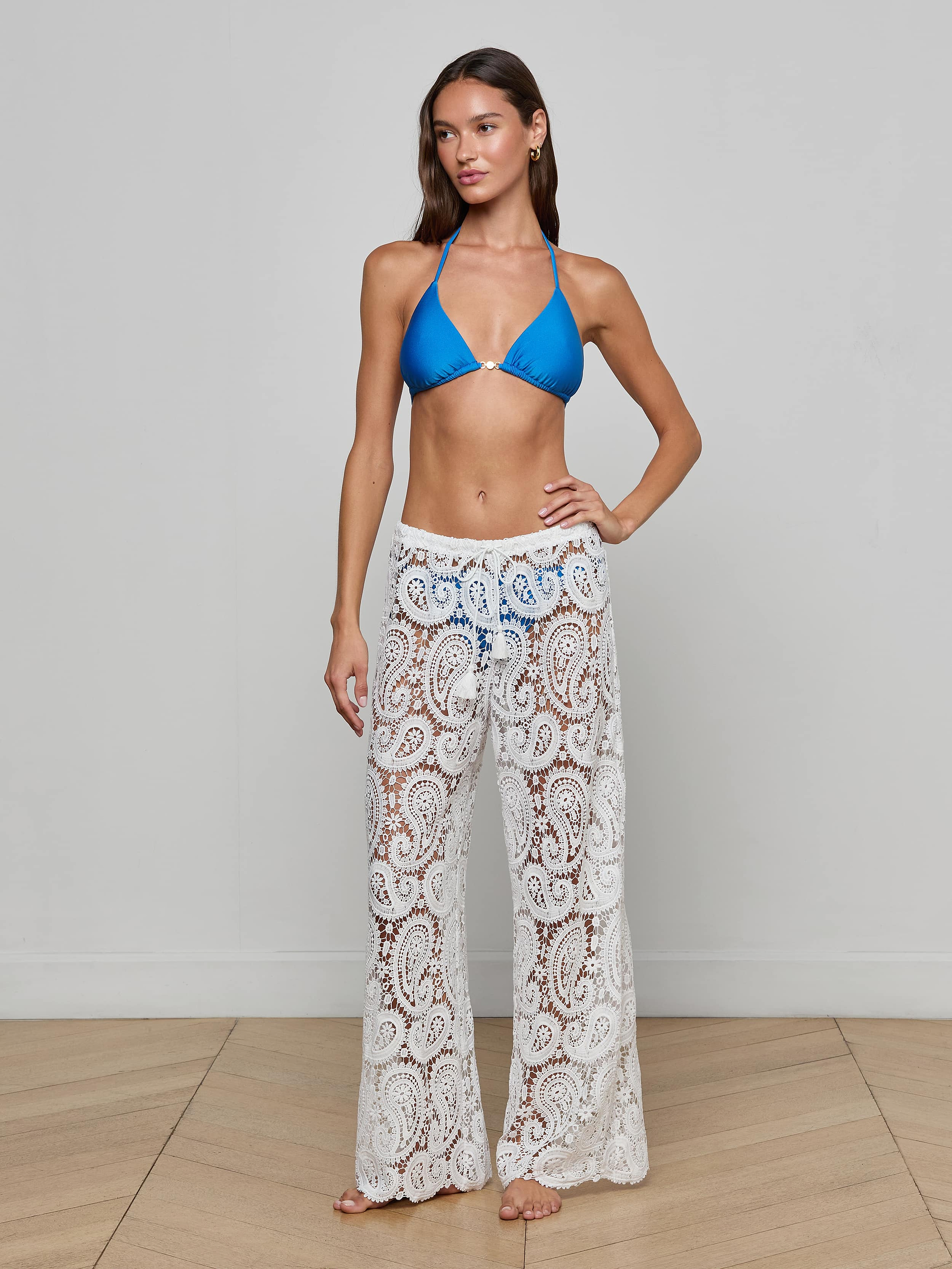 A woman stands barefoot against a plain light background, wearing a blue bikini top paired with the Geraldine Cover-Up Pant—white bohemian pants featuring a paisley crochet pattern.