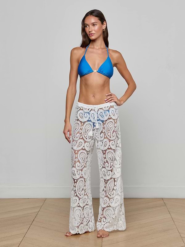 A woman with long brown hair poses barefoot on a wooden floor against a light gray wall, wearing the Geraldine Cover-Up Pant—white, sheer, and paisley crochet—for an effortlessly chic resort wear look.