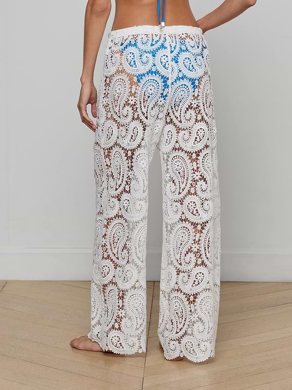 A person stands barefoot against a plain background, wearing the Geraldine Cover-Up Pant—a white, sheer, paisley crochet bohemian style—paired with a blue bikini top.