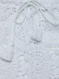 Close-up of the intricate white lace fabric of the Geraldine Cover-Up Pant, featuring two white tassels attached to thin cords resting on the lace—a perfect detail for resort wear or bohemian style.