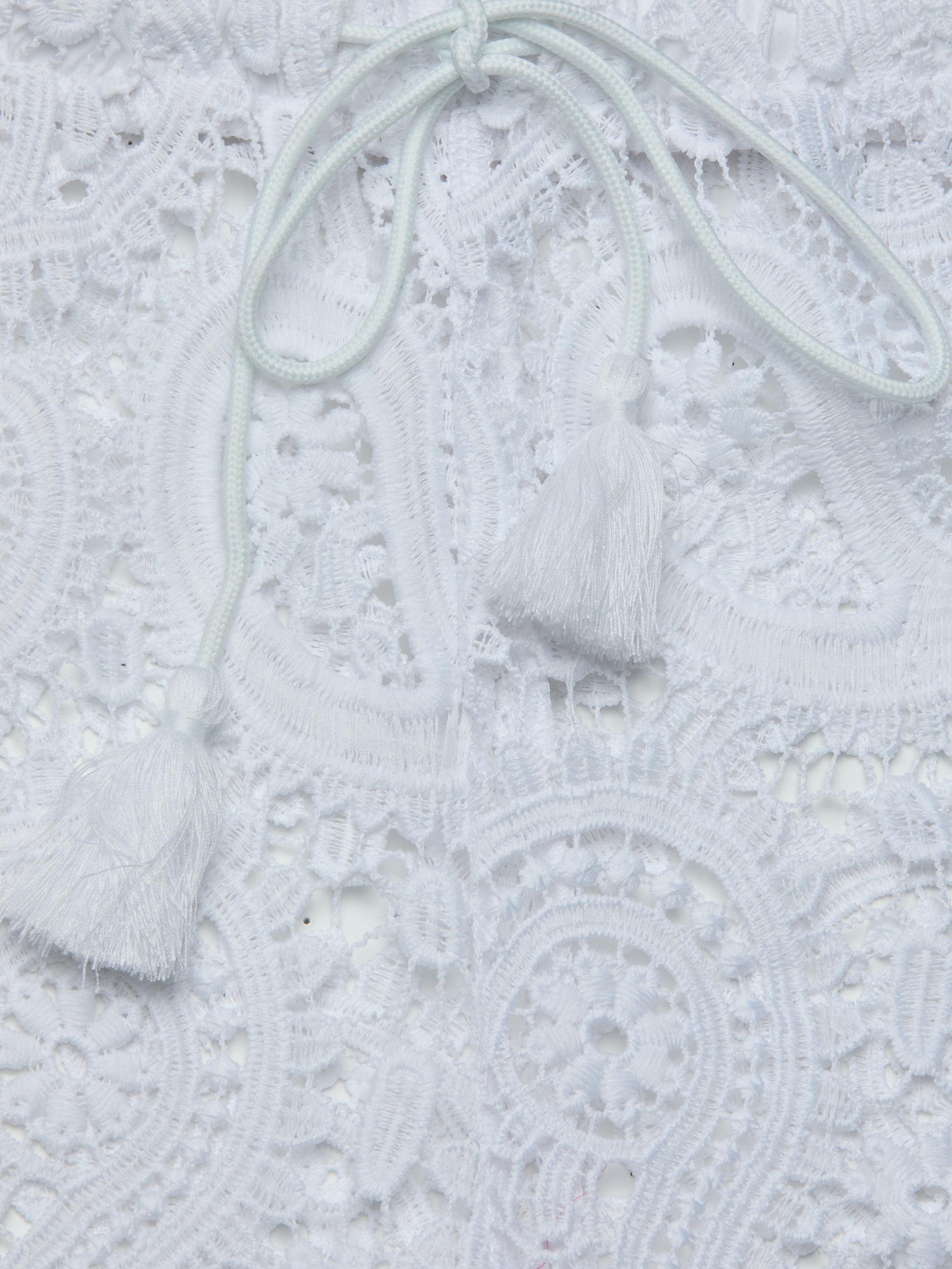 Close-up of the intricate white lace fabric of the Geraldine Cover-Up Pant, featuring two white tassels attached to thin cords resting on the lace—a perfect detail for resort wear or bohemian style.