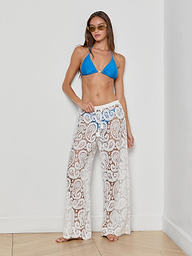 A woman stands confidently against a plain white background, wearing the Geraldine Cover-Up Pant—a sheer white, paisley crochet bohemian style—paired with a blue bikini top and sunglasses, with one hand on her hip.