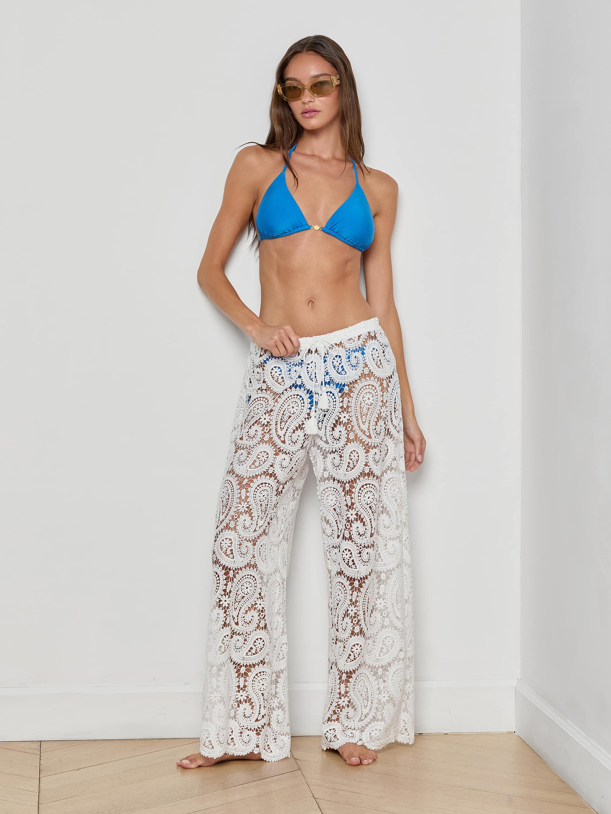 A woman stands confidently against a plain white background, wearing the Geraldine Cover-Up Pant—a sheer white, paisley crochet bohemian style—paired with a blue bikini top and sunglasses, with one hand on her hip.