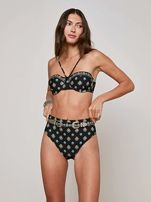 A woman with long brown hair wears the Jean Bikini Bottom, a high-waisted black geometric print piece, paired with a matching bandeau top. She stands against a plain light background, gazing to the side with a neutral expression.