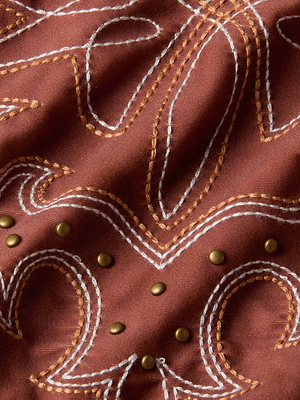 Close-up of the Lisa Plunge One-Piece Swimsuit by L'AGENCE Sale, showing brown suede-look fabric with white and tan Western-style embroidery, swirling designs, and small gold metallic studs arranged in a curved line.