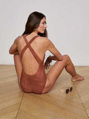 A woman with long brown hair sits on a wooden floor, facing away, wearing the L'AGENCE Sale Lisa Plunge One-Piece Swimsuit with crossed straps and a suede-look finish. Sunglasses lie on the floor beside her right hand.
