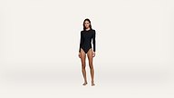 A woman stands barefoot against a plain white background, wearing a long-sleeved black bodysuit.