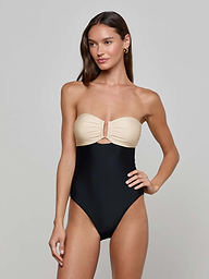 A woman with long brown hair poses against a plain light background, wearing the Emilia Bandeau One-Piece Swimsuit. The swimsuit features a beige bandeau-style top with ruched front detailing and a contrasting black high-leg bottom. She accessorizes her look with gold hoop earrings.