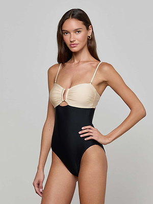 A woman with long brown hair models the L'AGENCE Emilia Bandeau One-Piece Swimsuit, featuring a beige top, black bottom, thin straps, ruched bust, and front cutout, against a plain gray background.