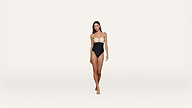 A woman stands barefoot against a plain white background, wearing a strapless one-piece swimsuit with a beige top and black bottom.