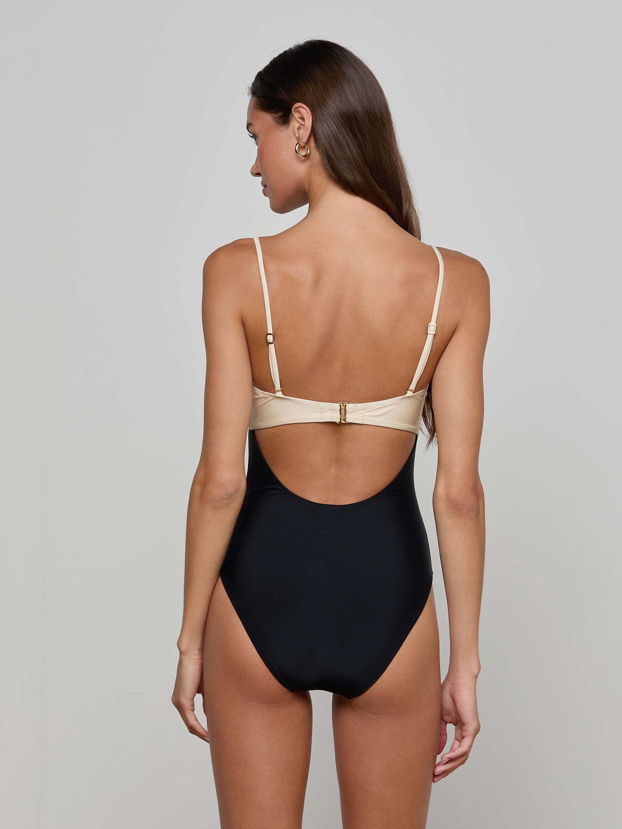 A woman with long brown hair wears the Emilia Bandeau One-Piece Swimsuit, a color-blocked black and beige bandeau style with a high-leg cut, standing with her back to the camera against a plain light background.