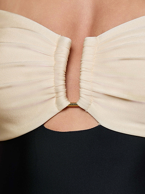 Close-up of the L'AGENCE Emilia Bandeau One-Piece Swimsuit, a strapless cream ruched top with a gold metal accent and small keyhole cutout below the bust, styled over a black garment.