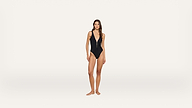 A woman stands barefoot wearing a black one-piece swimsuit with a deep V-neck, facing forward against a plain white background.