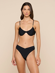 A woman stands facing forward, wearing the Tenali Bikini Top, an underwire black bra bikini top paired with high-waisted, V-cut bottoms. The background is plain and light-colored.