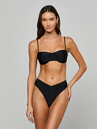 A woman stands against a light background wearing the Alexandria Bikini Top, which features an underwire balconette style, paired with matching high-waisted V-cut black bikini bottoms. She has long, straight hair and a relaxed expression.